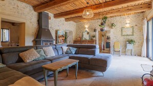 A cozy living room with a stone wall, wooden ceiling beams, a large sofa, a small table, and a hanging light fixture. - Guest Room “L’Aphyllanthe” with Large Shared Courtyard and Wi-Fi (Saint-Remèze)