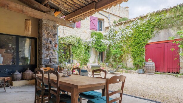 A rustic outdoor dining area with a wooden table and chairs, surrounded by stone walls and a red gate. - Guest Room “L’Aphyllanthe” with Large Shared Courtyard and Wi-Fi (Saint-Remèze)