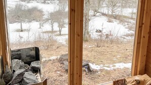 Blick durch ein Fenster in einen Raum mit Kamin aus Stein, Holzboden und Aussicht auf eine verschneite Landschaft mit Bäumen und einem Traktor.