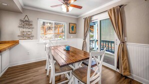 Un coin repas avec une table en bois et des chaises blanches, un ventilateur de plafond et une vue sur l’extérieur par une fenêtre.