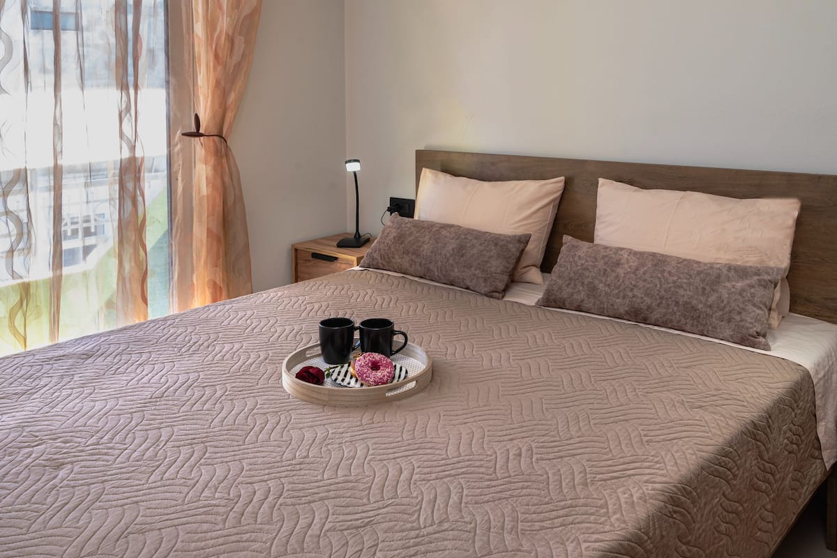 A neatly made bed with a tray of two mugs and a donut, a nightstand with a lamp, and a window with curtains.