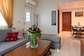Ein modernes Wohnzimmer mit einem grauen Sofa, einem Holztisch und einer Glasvase mit Blumen. Im Hintergrund ist eine Essecke mit Tisch und Stühlen zu sehen.