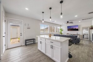 Een moderne keuken met witte kastjes, een centraal eiland, hanglampen en een televisietoestel met stadszicht.