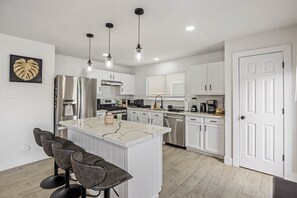 A modern kitchen with white cabinetry, a central island with bar stools, and pendant lighting. - Htown Getaway | Game Room BBQ Fire Pit Sleeps 16 (Houston)