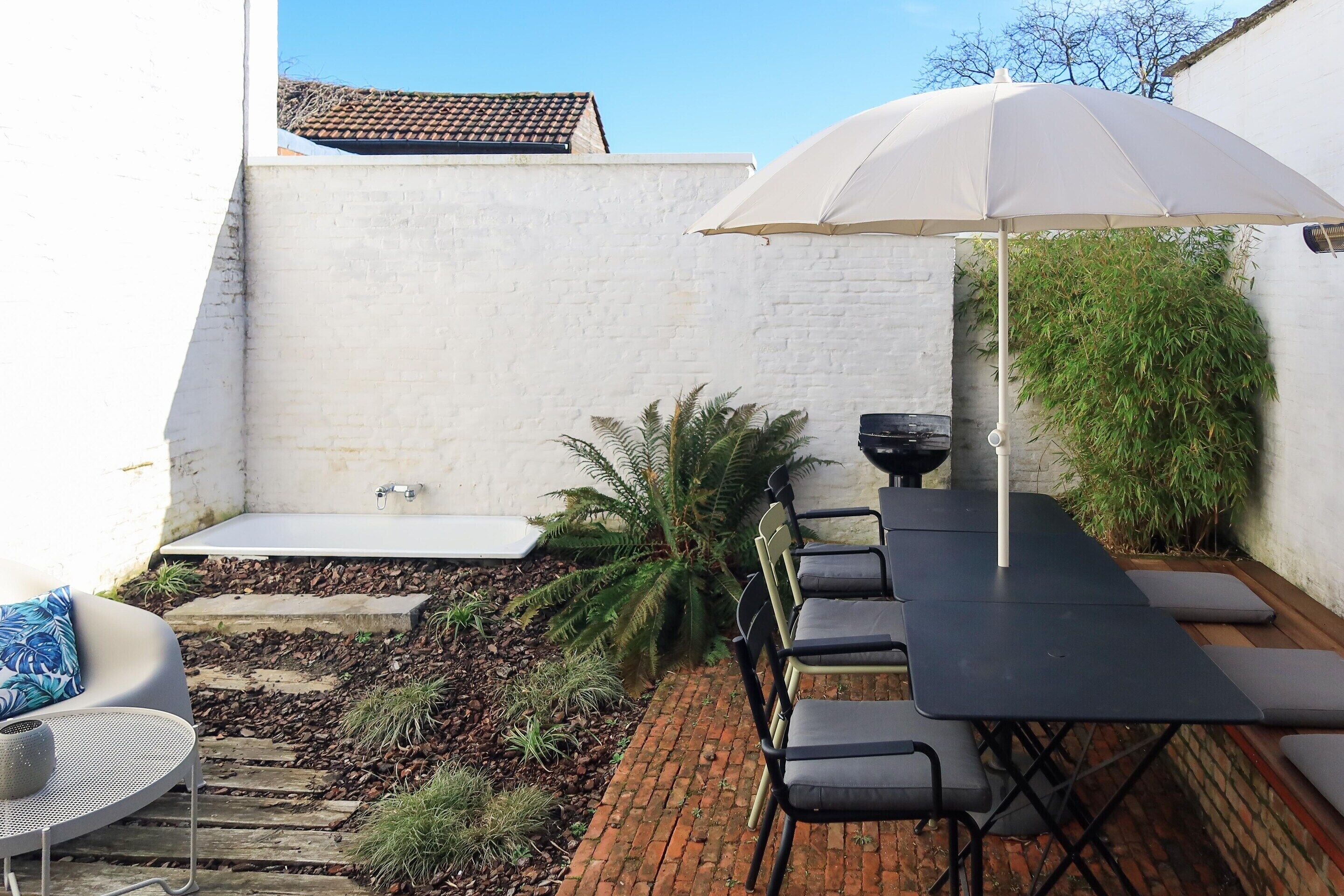 Un patio con una mesa y sillas, una sombrilla blanca, un jardín pequeño con plantas y un camino de ladrillos.