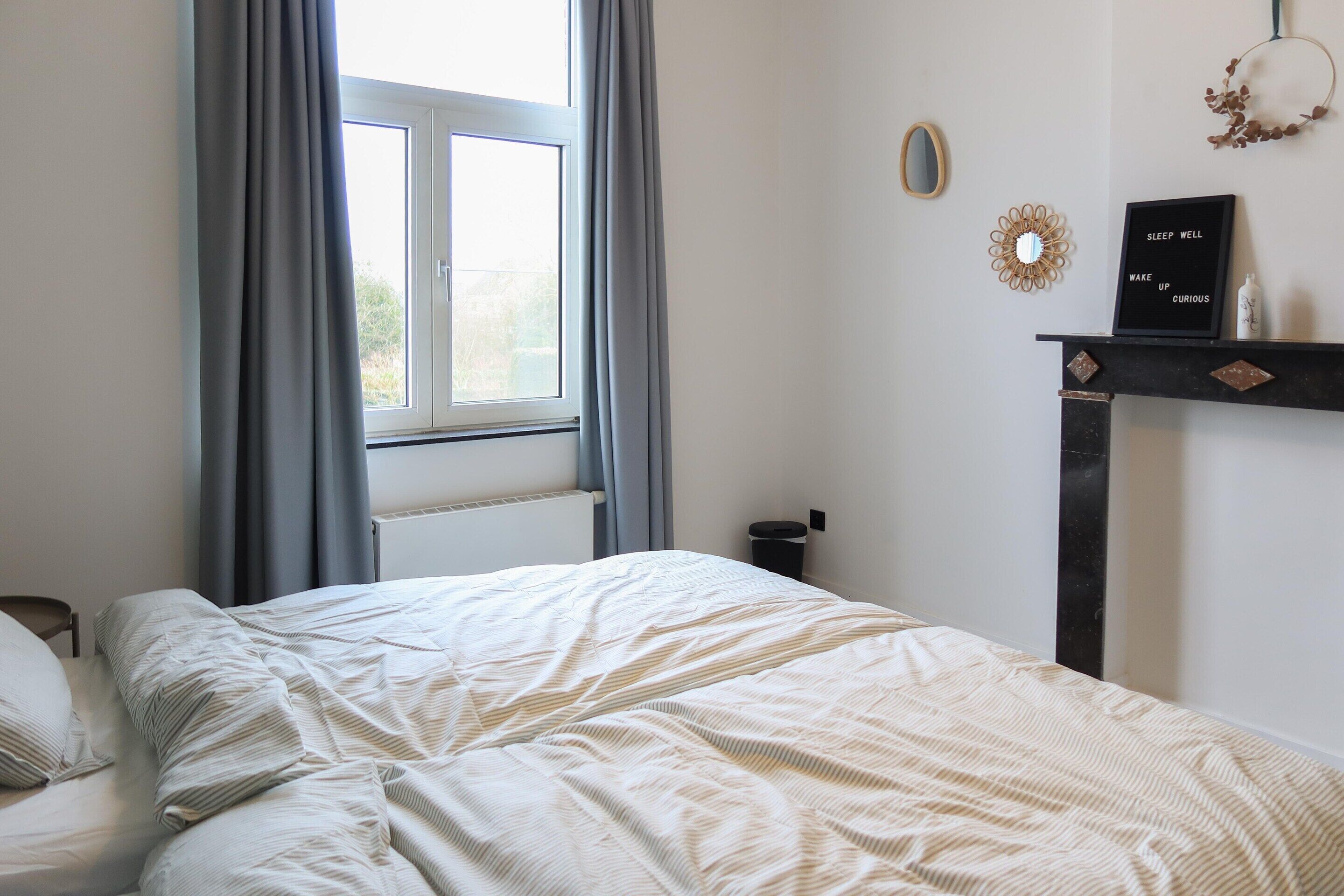 A bedroom with a bed, a window with curtains, a wall-mounted mirror, a decorative mirror, a framed motivational quote, and a small table with a bottle.
