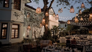 An outdoor dining area with tables set for a meal, surrounded by trees and a building with large windows. - The Manor (Colombo)