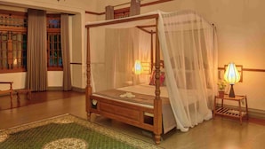 A four-poster bed with a canopy, a wooden floor, a chandelier, a bedside table with a lamp, and a view of a window with curtains. - The Manor (Colombo)