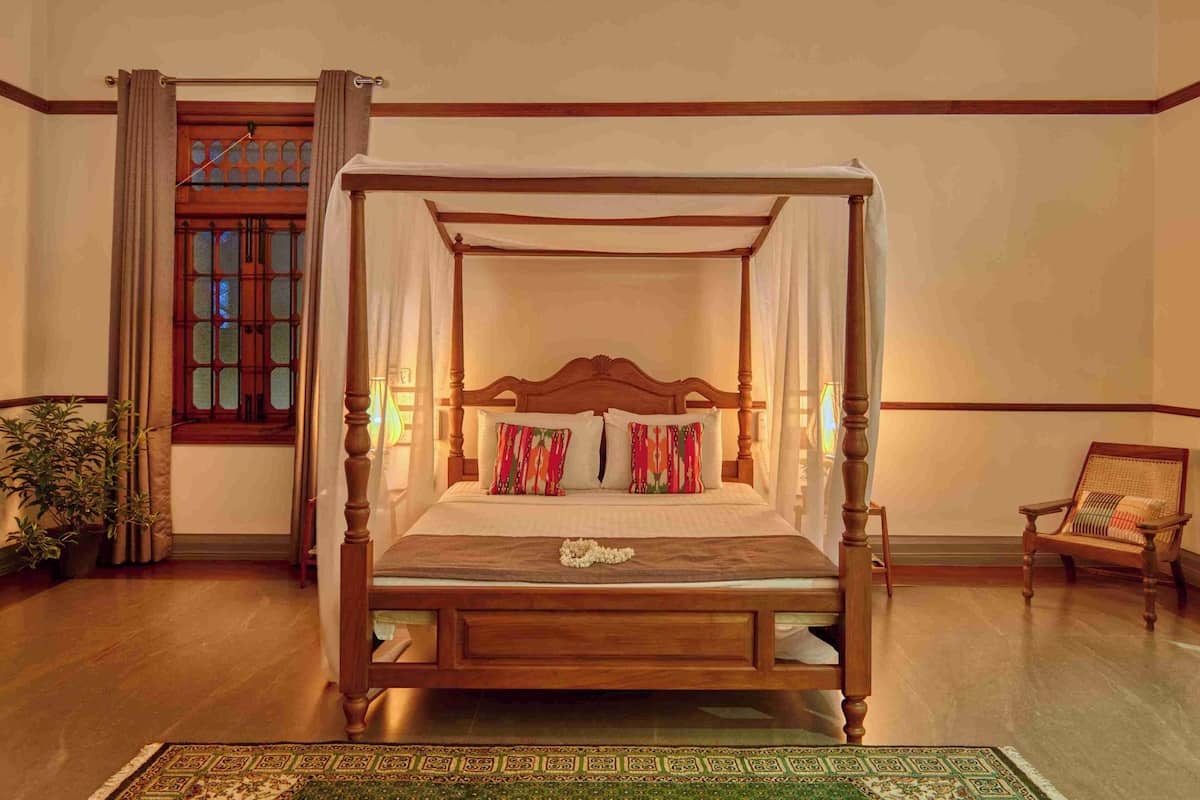 A four-poster bed with a canopy, a wooden chair, and a potted plant in a room with wooden flooring and a large window.