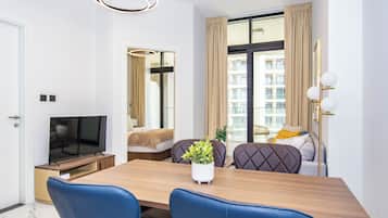 A modern hotel room with a wooden dining table, blue upholstered chairs, a flat-screen TV, a potted plant, and a view of the city through large windows.