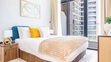 Een moderne hotelkamer met een groot bed, een nachtkastje, een raam met uitzicht op een gebouw en een kunstwerk in een lijst aan de muur.
