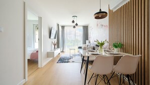 Un espace de repas moderne avec une table dressée pour deux personnes, un canapé en arrière-plan et une vue sur l’extérieur grâce à de grandes fenêtres.