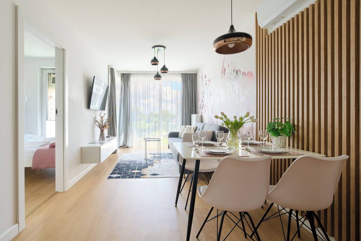 Un comedor moderno con una mesa preparada para dos, un sofá al fondo y vistas del exterior a través de amplios ventanales.