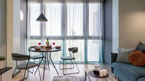 A modern living room with a dining area, large windows with sheer curtains, a hanging light fixture, and a sofa with cushions. - 2-Room Apart near the Botanical Garden (Minsk)