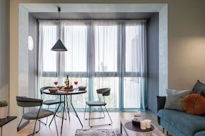 Een moderne woonkamer met een eethoek, grote ramen voorzien van dunne gordijnen, een hanglamp en een bank met kussens.