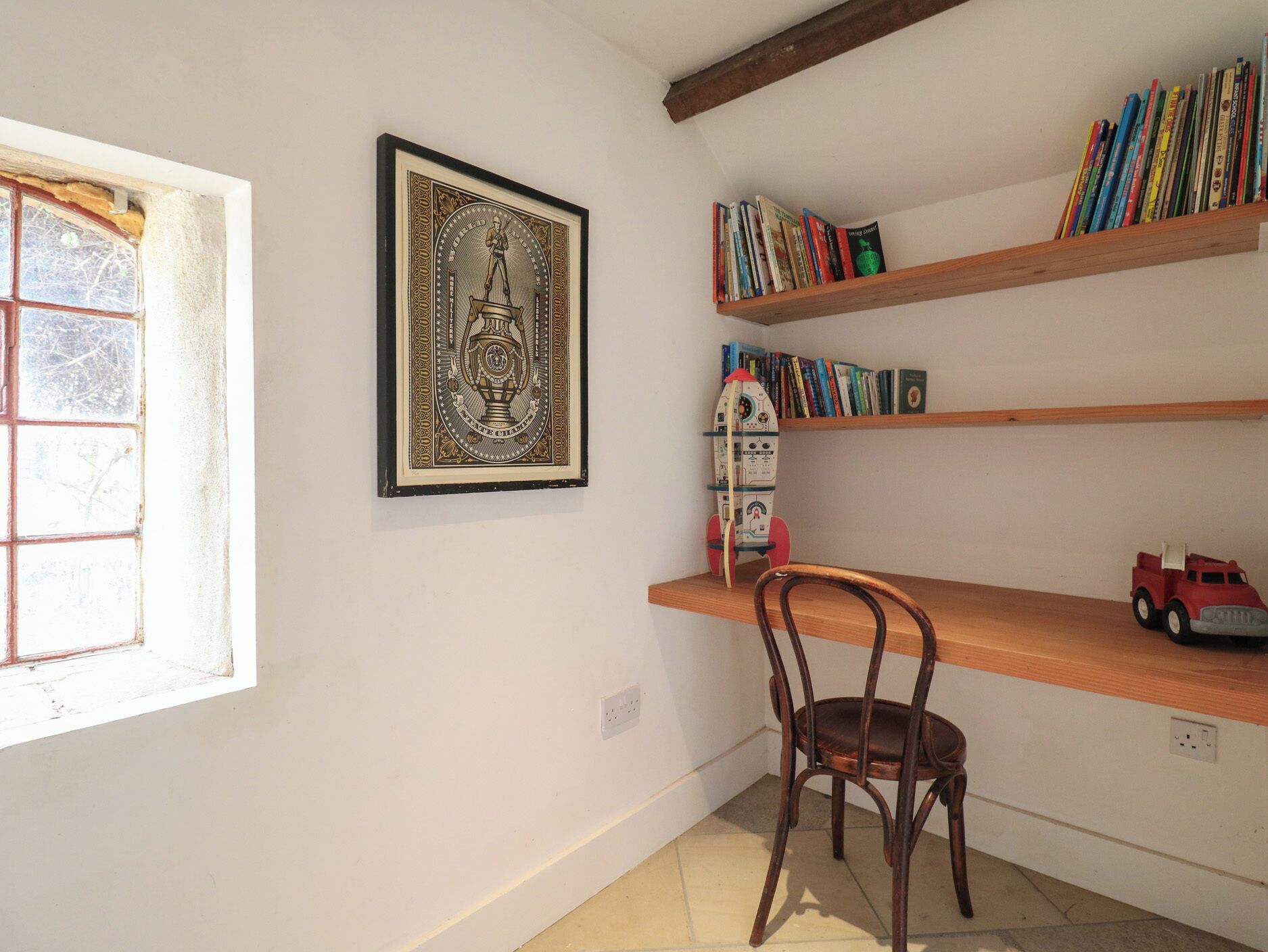 A room with a wooden chair, a bookshelf with books, a framed picture, and a window with a view of trees.