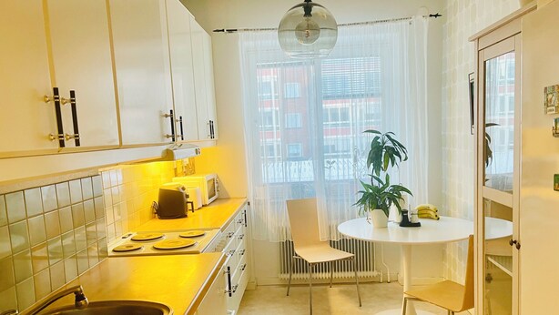 Une cuisine compacte équipée d’un évier, d’une cuisinière et de meubles blancs. On y trouve un coin repas avec une table et des chaises, ainsi qu’une plante en pot posée sur la table.