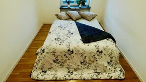 Une petite pièce étroite, avec un lit, deux plantes en pot et une fenêtre donnant sur des immeubles.