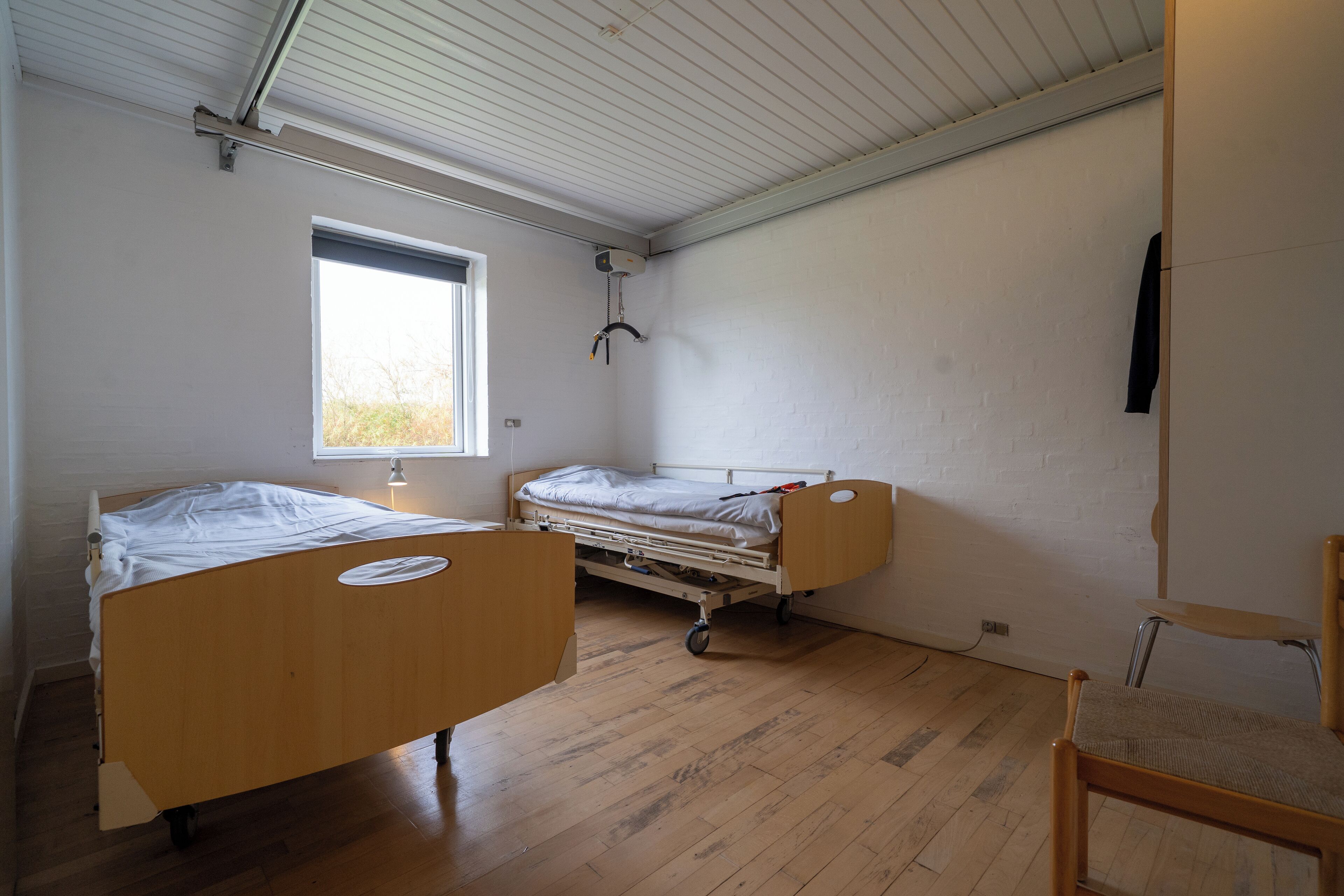 A room with two wooden beds, a window, and a chair.