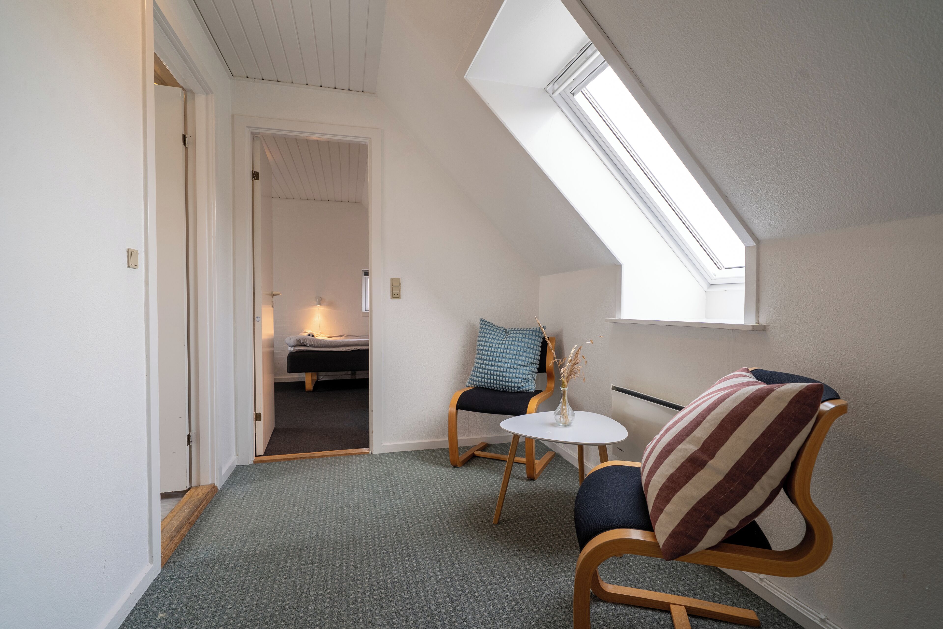 Une pièce chaleureuse avec un plafond en pente, une petite table ronde, deux chaises et un lit à l’arrière-plan.