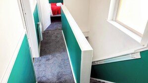 Un pasillo estrecho con paredes verde mar, una escalera con alfombra y una cama pequeña con una manta roja.