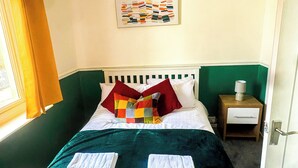Una cama bien hecha con un colcha colorida, almohadas blancas y dos toallas sobre la cama. Hay una mesita de noche con una lámpara y un cuadro enmarcado en la pared sobre la cama.