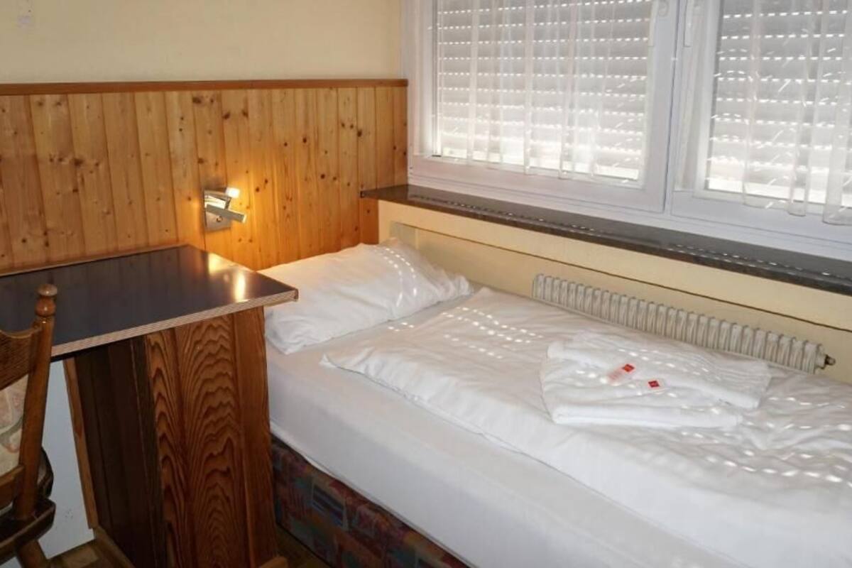 A small, cozy room with a wooden headboard, a single bed with white bedding, a small table, and a window with blinds.