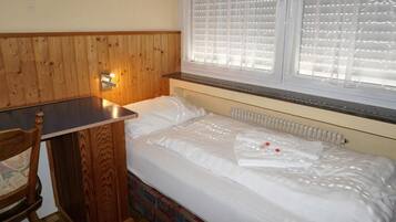 Ein kleiner, gemütlicher Raum mit einem Holzkopfteil, einem Einzelbett mit weißer Bettwäsche, einem kleinen Tisch und einem Fenster mit Jalousien.