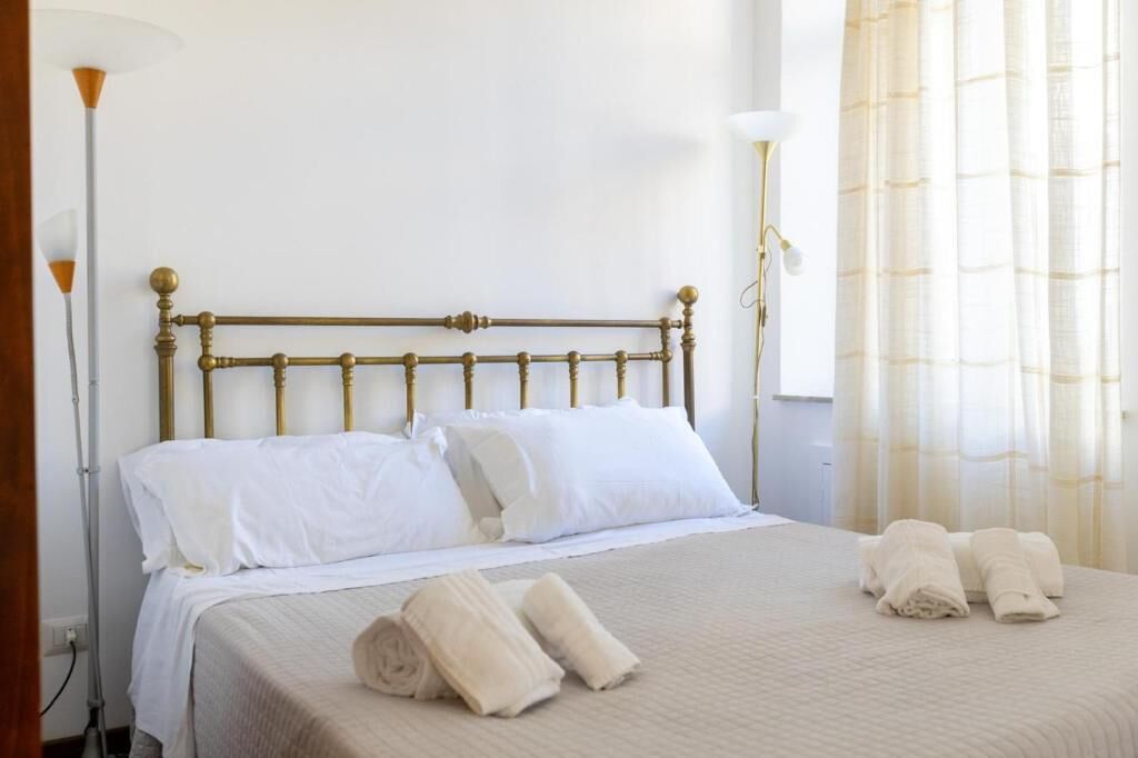 A neatly made bed with white linens and a gold headboard, a bedside lamp, and rolled towels on the bed.