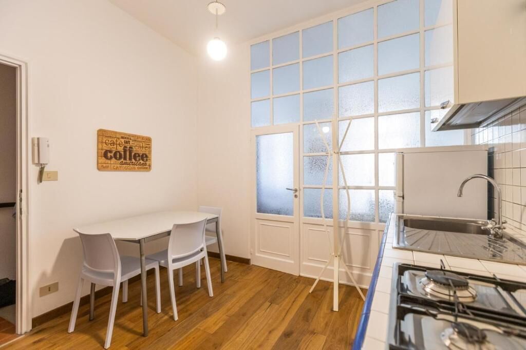 Una pequeña cocina con estufa a gas, un lavamanos y una ventana con vidrio esmerilado. Hay una mesa blanca con dos sillas y un cartel con motivos de café en la pared.