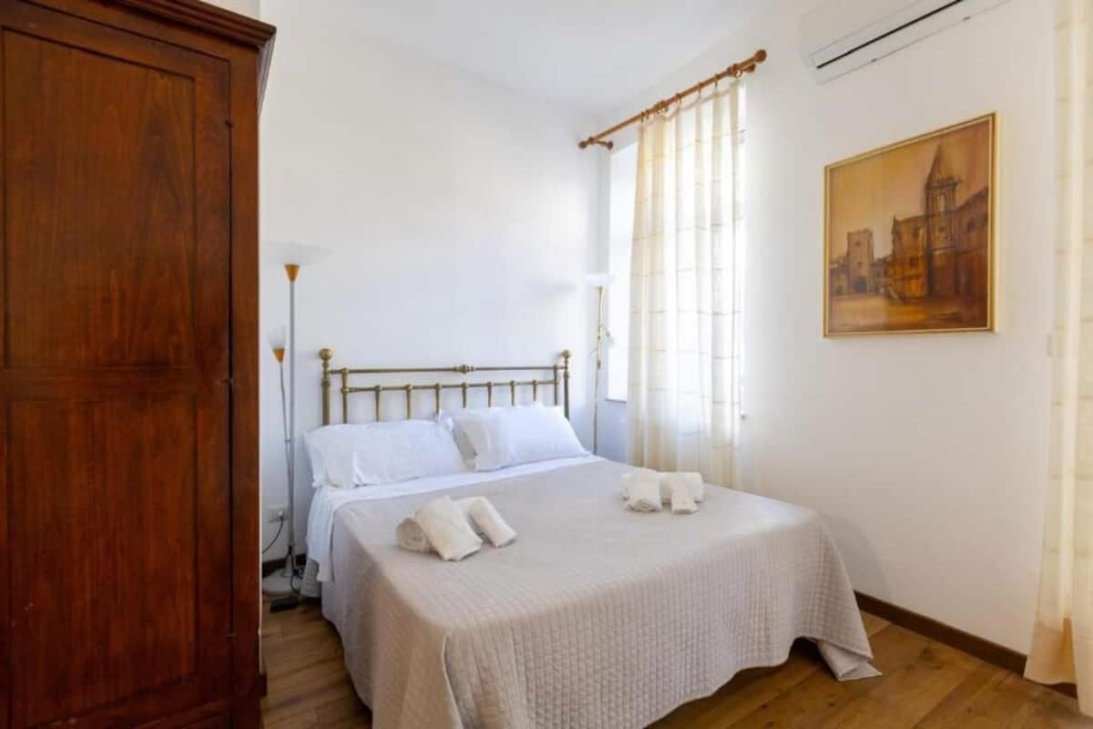 A bedroom with a wooden wardrobe, a bed with white linens, a painting on the wall, and a window with sheer curtains.