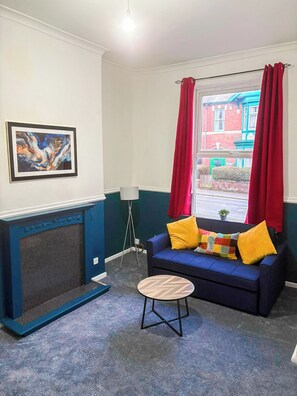 A living room with a blue sofa, a small round table, a framed abstract painting, and a window with red curtains. - Cozy Apt Near ASDA & Molineux • On-Site Parking (Wolverhampton)