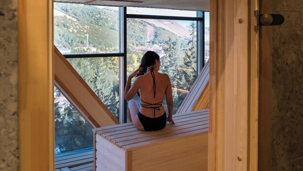 Una persona seduta su una panca di legno in una stanza con ampie finestre, con vista su un paesaggio montuoso.