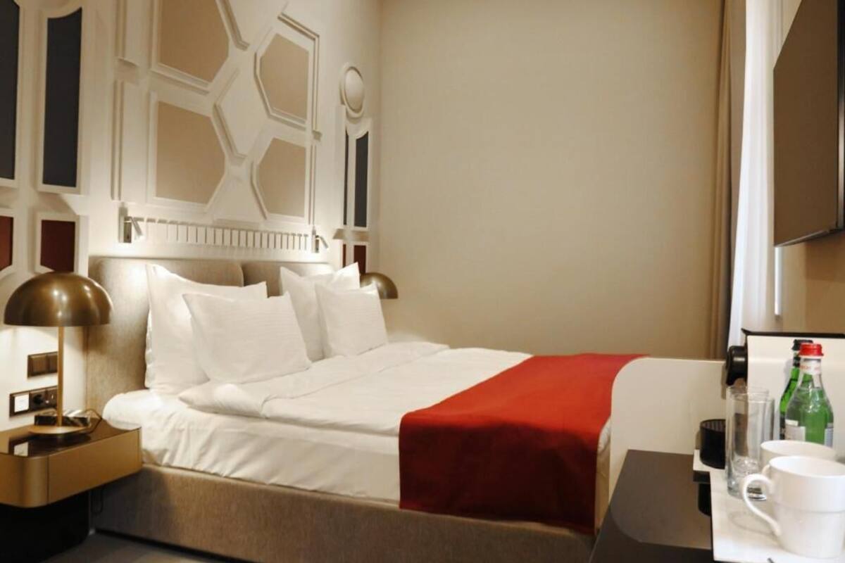 Habitación de hotel con una cama grande, una mesita de noche, una manta roja, una lámpara y un minibar.