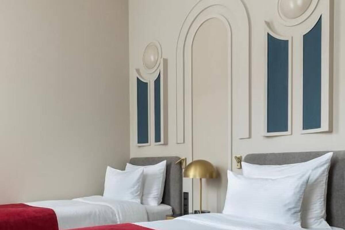 Habitación de hotel con dos camas, una colcha roja y blanca, una lámpara dorada y una pared con paneles decorativos.