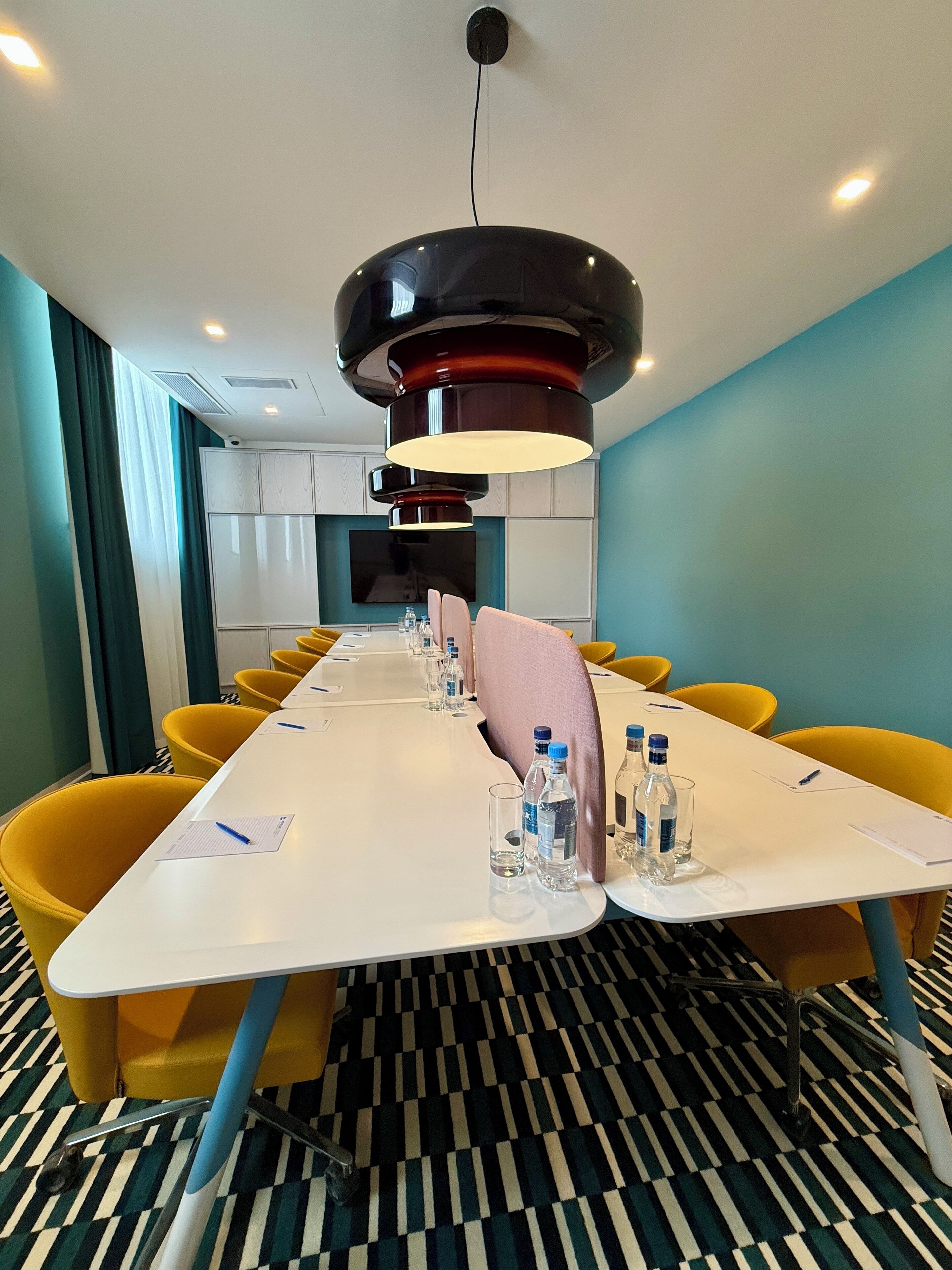 A conference room with a long table, yellow chairs, water bottles, and a large pendant light.