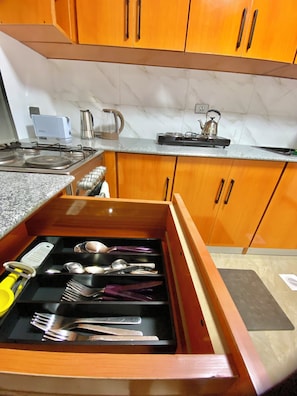 A kitchen with wooden cabinets, a countertop, a stove, a kettle, and a drawer with utensils.