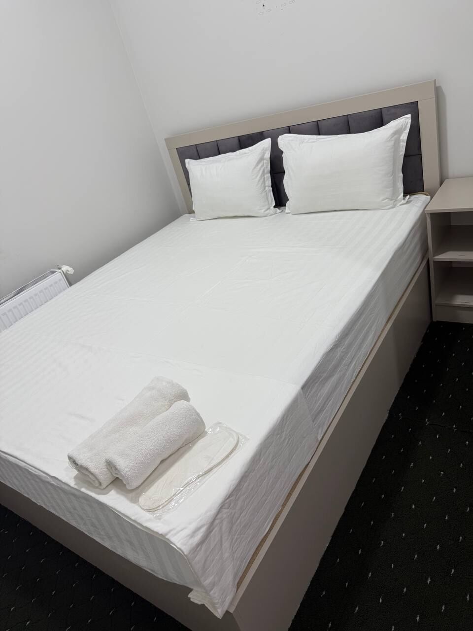 Un letto rifatto con lenzuola e cuscini bianchi, un comodino e un termosifone.