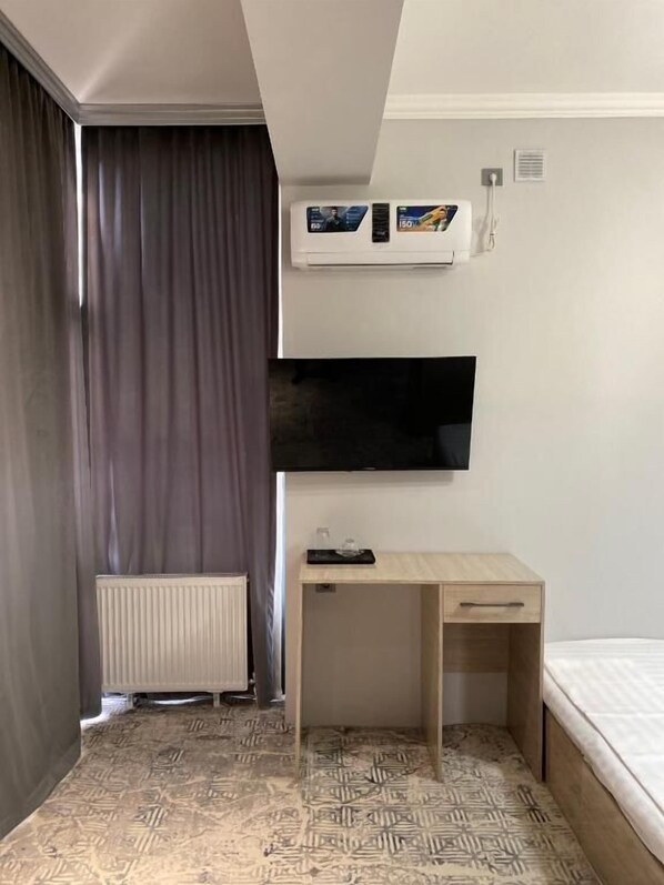 Et hotelværelse med seng, skrivebord, fjernsyn ophængt på væggen, en radiator og et gulv med mønster.