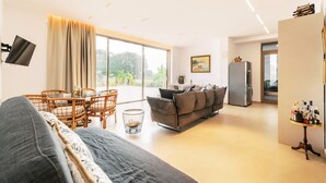 Una sala de estar moderna con sofá, mesa de comedor y sillas, una zona de cocina con refrigerador, y un ventanal con vistas a zonas verdes.