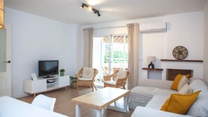 En moderne stue med et fladskærms-tv, en hvid sofa, to fletstole, et træsofabord og en skydedør, der fører ud til en balkon.
