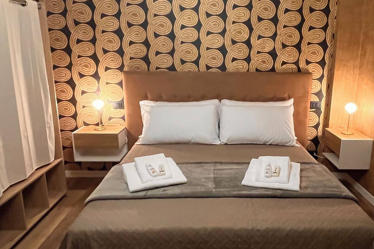 A hotel room with a patterned wallpaper, a large bed with two pillows, two bedside tables with lamps, and a neatly made bed with towels and a remote control.