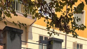 Ein verschlossener Eingang mit einem Metallgitter und einem Betonpfeiler, einem Briefkasten sowie ein Gebäude mit gelber Fassade im Hintergrund.