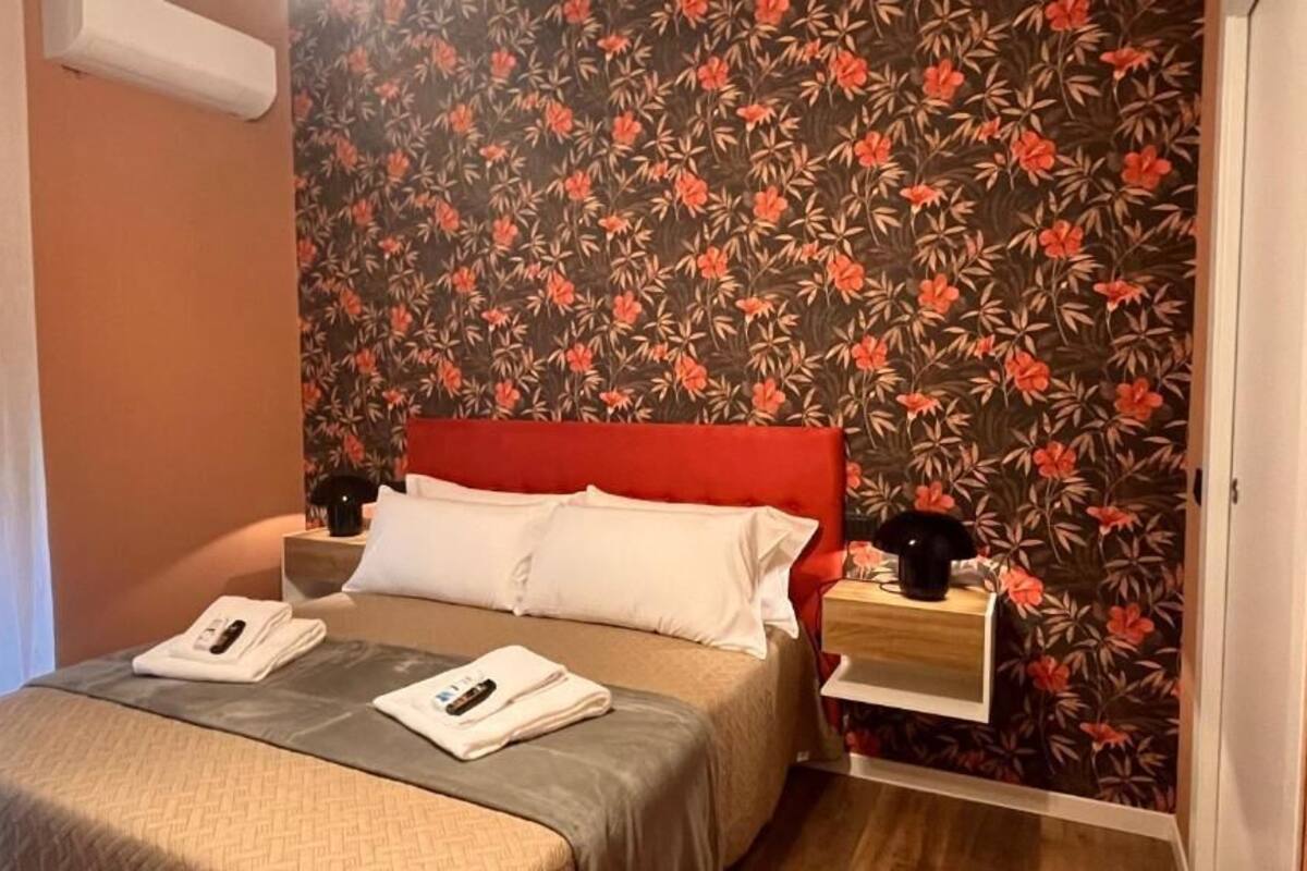 A hotel room with a bed, a nightstand, a bedside table, and a floral wallpaper.
