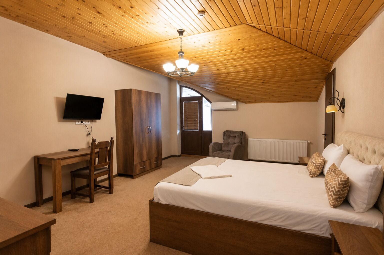Quarto com teto de madeira, uma cama, escrivaninha com cadeira, televisão e guarda-roupa.