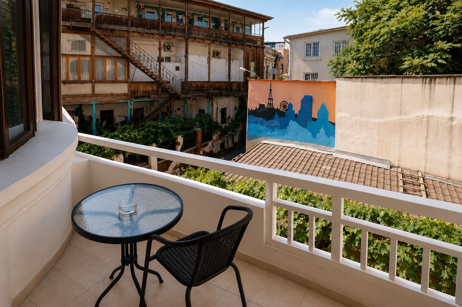 Un balcón con una mesa de vidrio y dos sillas, con vista a un patio con edificios y un mural.