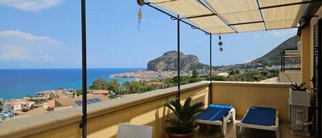 A balcony with a view of the sea, a table with a floral tablecloth, two sun loungers, and a potted plant.