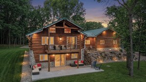 A two-story log cabin with a balcony, stone foundation, and outdoor seating area.