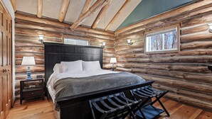A log cabin bedroom with a bed, two bedside tables with lamps, a bench, and a window with a view of trees.
