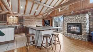 A log cabin interior with a stone fireplace, wooden bar stools, a kitchen island, and a large window.
